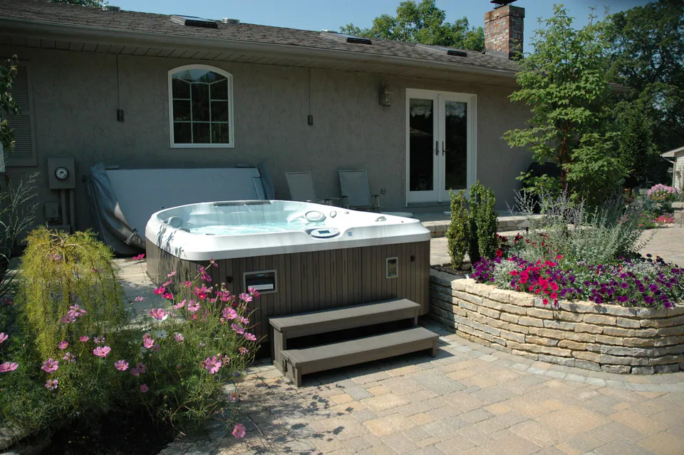 Hot tub set on a paver patio with steps, a raised stone planter and flowering plants